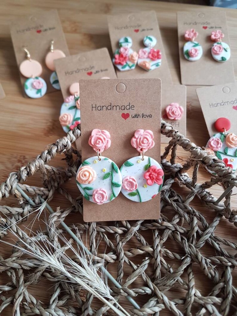 May include: A pair of round white earrings featuring raised pink and peach floral designs with green leaves and small yellow dots. The earrings are attached to a brown cardboard display card with the text Handmade with love printed on it.