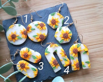 May include: Four pairs of dangle earrings featuring white bases with yellow sunflower designs and green leaves. Pair 1 is circular, pair 2 is hexagonal, pair 3 is oval, and pair 4 is arch-shaped. Each earring has a gold-toned metal hook.