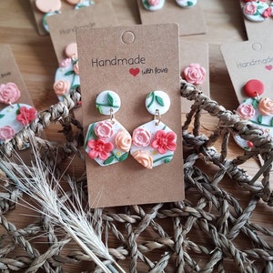 May include: A pair of white hexagonal polymer clay earrings featuring raised pink and peach floral designs with green leaves and stems. The earrings are attached to a brown cardboard display card printed with the text Handmade with love.