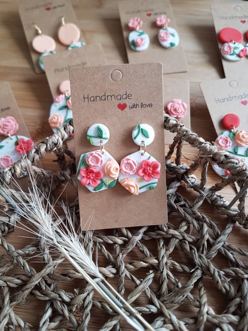 May include: A pair of white hexagonal polymer clay earrings featuring raised pink and peach floral designs with green leaves and stems. The earrings are attached to a brown cardboard display card printed with the text Handmade with love.