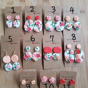 May include: Eleven sets of handmade polymer clay earrings displayed on brown cardboard cards. The designs feature floral motifs with pink, red, and peach roses, green leaves, and white backgrounds in various shapes like circles, ovals, and diamonds.