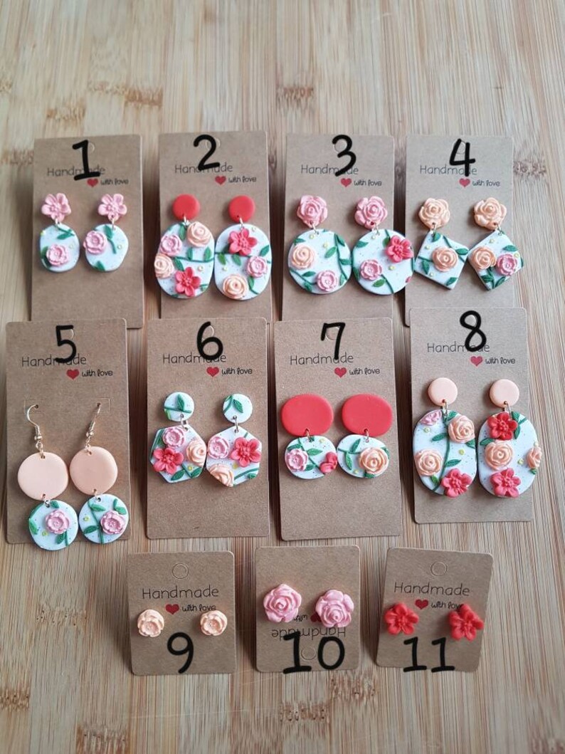 May include: Eleven sets of handmade polymer clay earrings displayed on brown cardboard cards. The designs feature floral motifs with pink, red, and peach roses, green leaves, and white backgrounds in various shapes like circles, ovals, and diamonds.