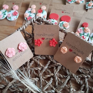 May include: A collection of handmade floral earrings displayed on brown cardboard tags. The earrings feature various sculpted flower designs in shades of pink, peach, and red, some with green leaf accents, attached to round or stud-style backings.