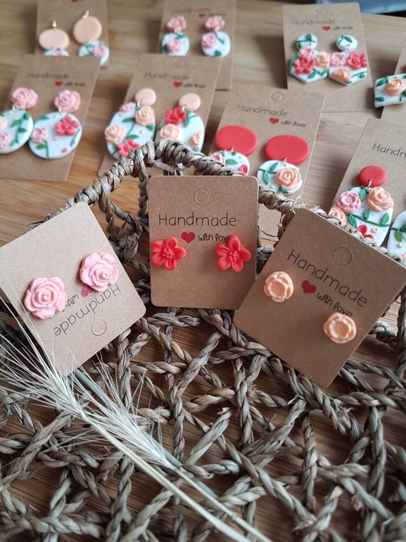 May include: A collection of handmade floral earrings displayed on brown cardboard tags. The earrings feature various sculpted flower designs in shades of pink, peach, and red, some with green leaf accents, attached to round or stud-style backings.