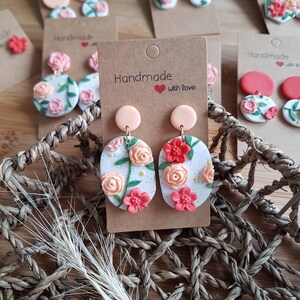 May include: Oval-shaped dangle earrings featuring a white base with speckled details and sculpted floral motifs in shades of peach, coral, and red, accented with green leaves. Each earring is attached to a solid peach circular stud on a card.