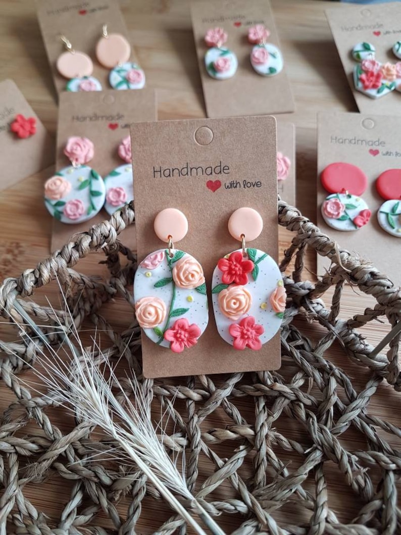 May include: Oval-shaped dangle earrings featuring a white base with speckled details and sculpted floral motifs in shades of peach, coral, and red, accented with green leaves. Each earring is attached to a solid peach circular stud on a card.