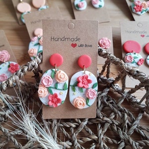 May include: A pair of oval-shaped earrings featuring a white base with speckled details, adorned with sculpted pink and peach-colored flowers and green vines. Each earring has a solid red circular stud top and is attached to a brown cardboard display card.