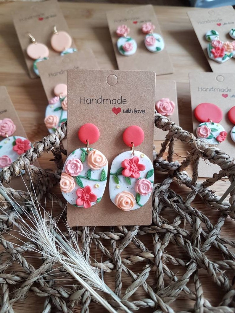 May include: A pair of oval-shaped earrings featuring a white base with speckled details, adorned with sculpted pink and peach-colored flowers and green vines. Each earring has a solid red circular stud top and is attached to a brown cardboard display card.