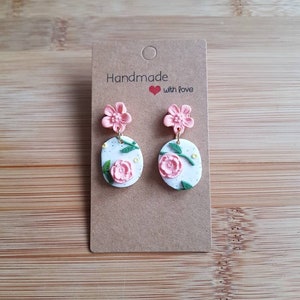 May include: A pair of dangle earrings on a brown card labeled Handmade with love. Each earring features a pink flower stud attached to a white oval pendant decorated with a pink floral motif, green leaves, and small yellow dots, connected by gold rings.