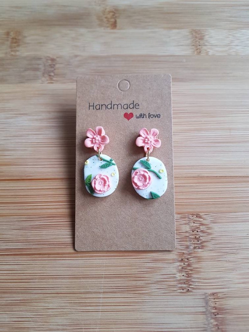 May include: A pair of dangle earrings on a brown card labeled Handmade with love. Each earring features a pink flower stud attached to a white oval pendant decorated with a pink floral motif, green leaves, and small yellow dots, connected by gold rings.
