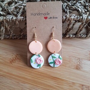 May include: Dangle earrings featuring two stacked circular charms. The top charm is solid peach, while the bottom charm is white with a speckled texture, a pink floral relief, and green stems. The earrings hang from gold-toned metal hooks on a brown card.