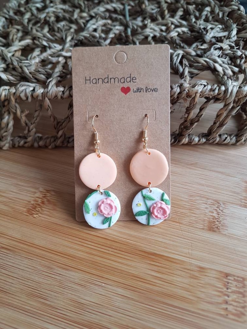 May include: Dangle earrings featuring two stacked circular charms. The top charm is solid peach, while the bottom charm is white with a speckled texture, a pink floral relief, and green stems. The earrings hang from gold-toned metal hooks on a brown card.
