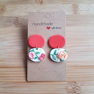 May include: A pair of dangle earrings on a brown cardboard display card labeled Handmade with love. Each earring features a solid red circular top piece connected to a white circular base with a speckled texture and a raised floral design.