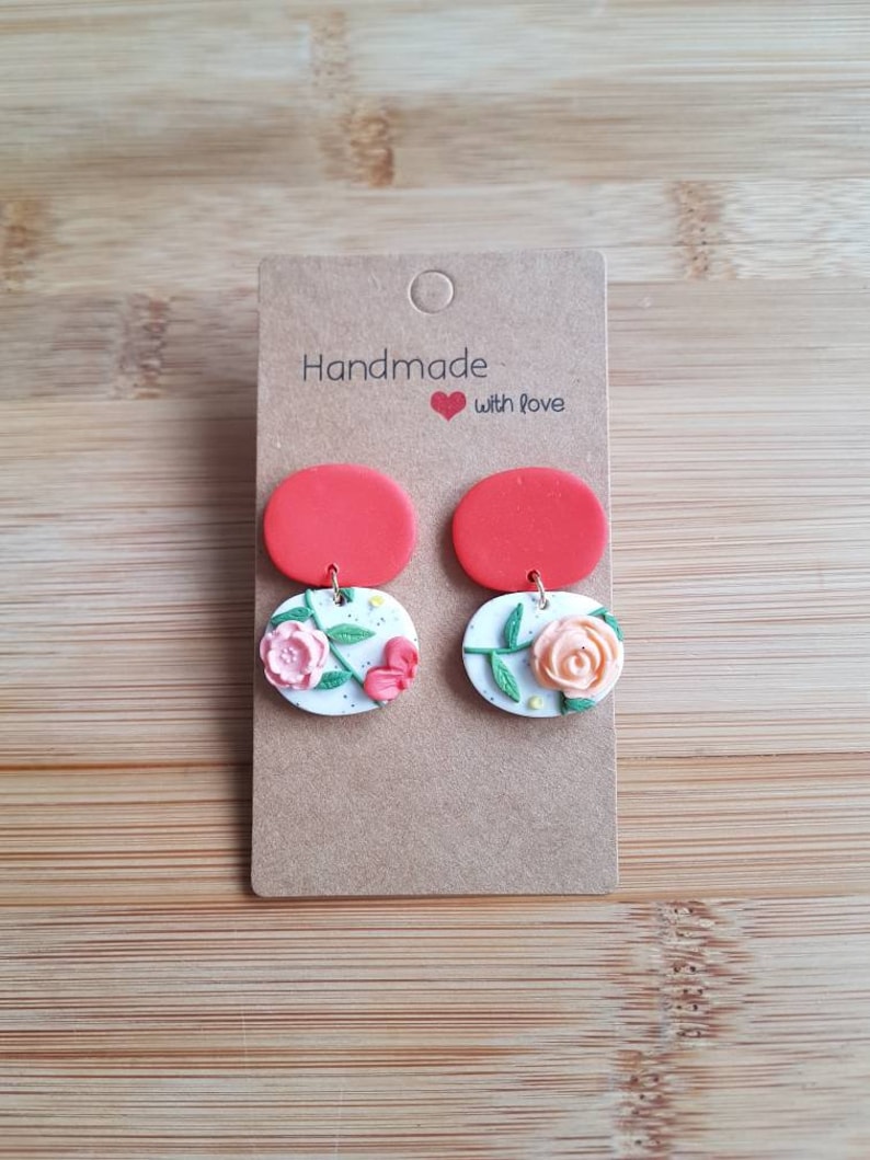 May include: A pair of dangle earrings on a brown cardboard display card labeled Handmade with love. Each earring features a solid red circular top piece connected to a white circular base with a speckled texture and a raised floral design.
