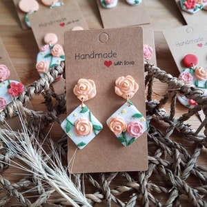 May include: Dangle earrings featuring peach-colored rose studs attached to white diamond-shaped pendants. The pendants display hand-sculpted peach and pink roses with green leaves and yellow dots on a speckled white background, mounted on brown cardstock.