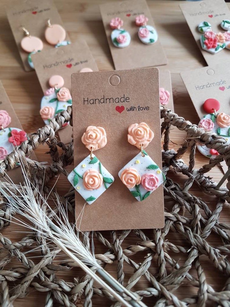 May include: Dangle earrings featuring peach-colored rose studs attached to white diamond-shaped pendants. The pendants display hand-sculpted peach and pink roses with green leaves and yellow dots on a speckled white background, mounted on brown cardstock.