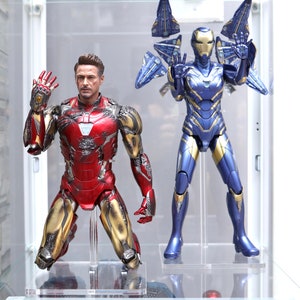 Puede incluir: Dos figuras de acci&oacute;n, una roja y dorada, la otra azul y dorada, est&aacute;n colocadas en soportes de pl&aacute;stico transparente. La figura roja y dorada est&aacute; arrodillada con un brazo levantado. La figura azul y dorada est&aacute; de pie con los dos brazos levantados. Las figuras se exhiben en una vitrina de vidrio.