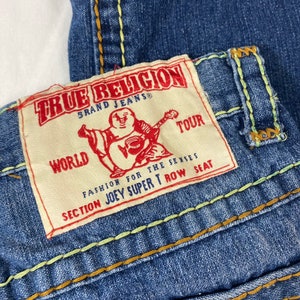 May include: A close-up of a blue denim jean with a True Religion brand label. The label features a white background with red text and a cartoon Buddha playing a guitar. The text on the label reads "True Religion Brand Jeans", "World Tour", "Fashion for the Senses", "Section Joey Super T Row Seat".