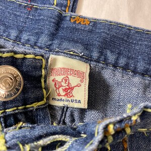 May include: Close-up of a blue denim jean with yellow stitching and a True Religion brand label. The label is white with red stitching and features a Buddha-like figure holding a guitar. The label reads "True Religion" and "Made in USA".