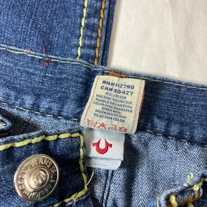 May include: A close-up of a blue denim jean with a yellow and orange stitching detail. The jean has a True Religion brand tag with the text "RN#112790 CA#30427 100% COTTON MACHINE WASH COLD TUMBLE DRY MEDIUM. COLOR MAY TRANSFER WHEN NEW WASH INSIDE OUT TO AETAIN COLOR."