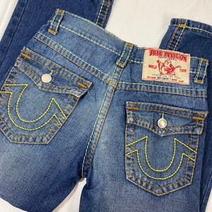 May include: Blue denim jeans with yellow stitching and a True Religion patch on the back pocket. The patch says "World Tour" and "Section Joey Upper T Row Seat".