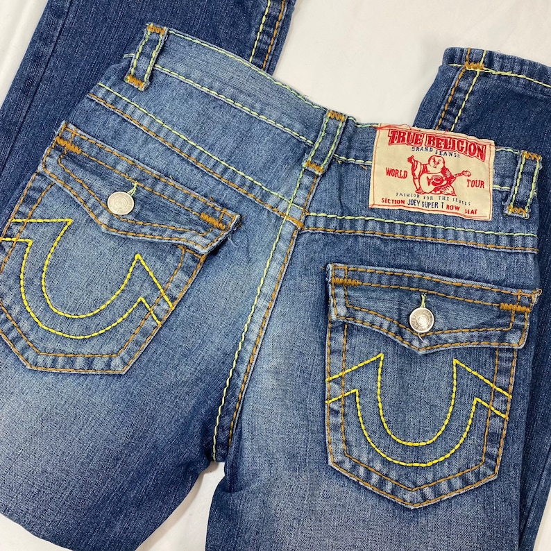 May include: Blue denim jeans with yellow stitching and a True Religion patch on the back pocket. The patch says "World Tour" and "Section Joey Upper T Row Seat".