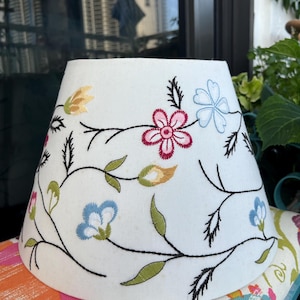 May include: A white tapered lampshade featuring an embroidered pattern of black vines with colorful flowers in shades of pink, light blue, and yellow, along with green leaves. The design is distributed across the surface of the fabric shade.
