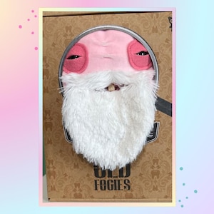May include: A plush toy character with pink skin, drooping eyelids, and a long white faux fur beard, packaged in a brown cardboard box labelled OLD FOGIES. The character is visible through a circular cutout in the front of the packaging.
