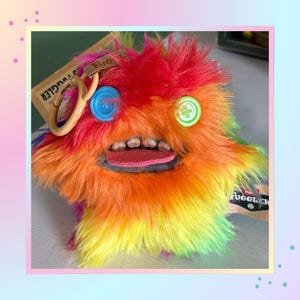 May include: A plush toy with long, rainbow-coloured faux fur, featuring a blue button eye on the left and a green button eye on the right. The creature has a wide mouth with visible human-like teeth, a pink tongue, and a tan plastic keychain ring.