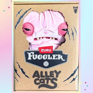 Op de afbeelding: Een Fuggler Alley Cats pluche speelgoed in een kartonnen doos. Het roze pluche speelgoed heeft rode accenten en zichtbare tanden. De doos heeft een uitgescheurd gedeelte dat het speelgoed onthult, met het Fuggler-logo en de tekst "Alley Cats".