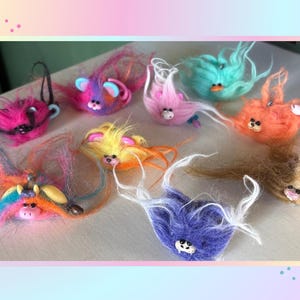 May include: A collection of nine small, fuzzy, multi-colored creatures with long, wispy hair. Each creature features a small, sculpted face with distinct ears, eyes, and noses in various bright colors including pink, purple, orange, yellow, and teal.