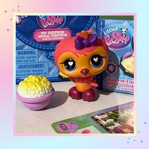 May include: A Littlest Pet Shop bird figurine with a pink head, orange body, and dark purple feet, featuring a purple bow on its head and heart-shaped pupils. Beside it sits a small purple bowl filled with yellow popcorn, with product packaging behind.