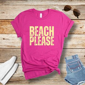 May include: A bright pink t-shirt with the words "BEACH PLEASE" in a yellow, distressed font. The shirt is folded on a wooden surface, with white sneakers, denim shorts, and sunglasses nearby. A casual summer outfit.