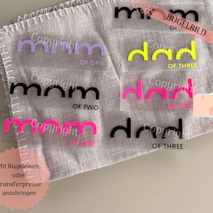 Può includere: Una collezione di disegni in vinile a trasferimento termico in vari colori, tra cui viola, nero, rosa e giallo. I disegni presentano le parole "mamma" e "papà" con il testo "Copyright" e "OF ONE, TWO, THREE". I disegni sono su un panno bianco.