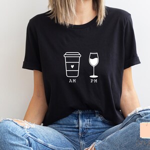 Puede incluir: Camiseta negra con un gráfico blanco de una taza de café y una copa de vino. El texto "AM" está debajo de la taza de café y "PM" debajo de la copa de vino. La camiseta es de material suave.