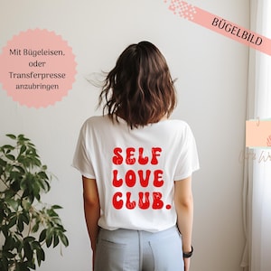 Può includere: Una maglietta bianca con le parole "SELF LOVE CLUB" in rosso, con lettere in stile retrò sul retro. La persona indossa pantaloni grigio chiaro. Un banner rosa con testo tedesco si trova nell'angolo in alto a destra.
