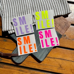 Può includere: Quattro adesivi quadrati con la scritta "SMILE!" in diversi colori neon: viola, giallo, arancione e rosa. Gli adesivi sono su un tagliere grigio scuro con manico in pelle, su una superficie di legno.