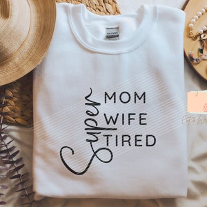 Op de afbeelding: Wit sweatshirt met de tekst "Super Mom Wife Tired" in zwarte letters. Het kledingstuk ligt opgevouwen op een licht oppervlak. Het merklabel is zichtbaar op de kraag.