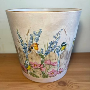 May include: A white ceramic planter featuring a painted design of a picket fence, flowers, and birds. The birds are blue and yellow, and the flowers are pink and white.