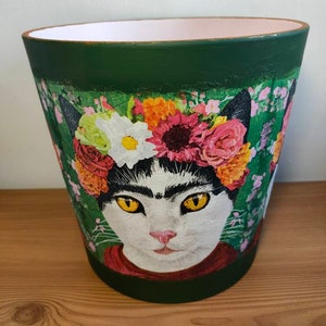 May include: Green ceramic planter with a floral design featuring a cat wearing a flower crown. The cat has black and white fur and yellow eyes.