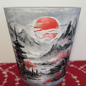 May include: A hand-painted ceramic pot with a Japanese landscape design. The design features a red sun, mountains, a pagoda, and cherry blossoms in shades of red, grey, and black. The pot is cylindrical and has a textured surface.