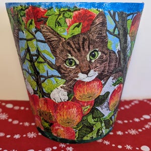 May include: A decorative flower pot with a design of a brown tabby cat surrounded by red apples and green foliage. The pot has a blue rim and sits on a red and white patterned surface. The cat has green eyes.