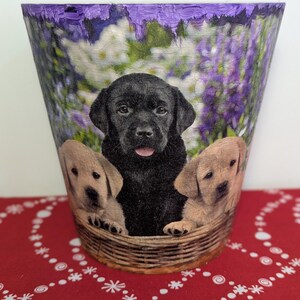 May include: A purple flower pot featuring a picture of three puppies. The puppies are a black labrador, a golden retriever, and a yellow labrador. The puppies are sitting in a wicker basket.