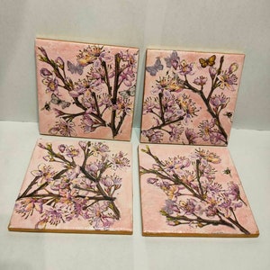 May include: Set of four square ceramic coasters with a pink background and a floral design featuring pink and white flowers, green leaves, and butterflies.