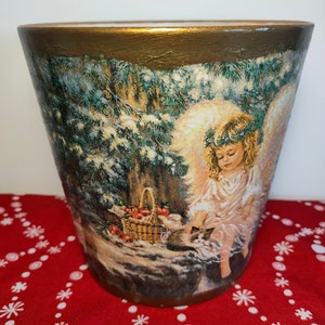 May include: A ceramic planter with a gold rim features a winter scene with a snow-covered forest, a small cat, and an angel with a basket of apples. The planter is decorated with a wintery scene.