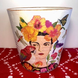 May include: White decorative planter with a portrait of a woman adorned with a floral crown, a parrot, and a butterfly. The planter has a textured surface and a gold rim. The design features vibrant orange, pink, and green floral and fauna elements.