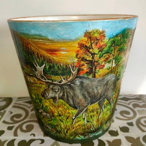 May include: A ceramic planter featuring a painted scene of a moose in a woodland setting. The background depicts a blue sky, green trees, and a yellow sunset.