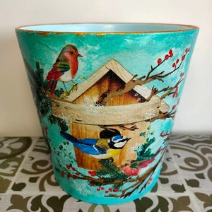 May include: A turquoise blue planter with a winter scene featuring a birdhouse, birds, and snow. The planter has a gold rim.