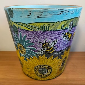 May include: A blue ceramic planter with a floral design featuring sunflowers, lavender, and bees. The planter has a light blue glaze and a gold rim.