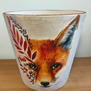 May include: A white ceramic planter featuring a painted image of a fox's face. The fox has orange fur, a black nose, and brown eyes. The planter is decorated with red and blue leaves and berries.